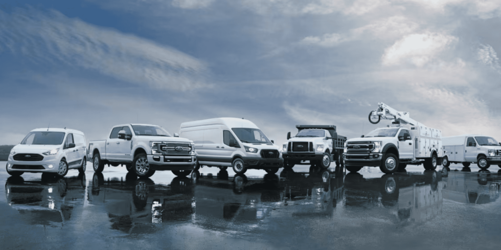 Fleet of six white commercial vehicles under cloudy skies reflecting on wet ground.